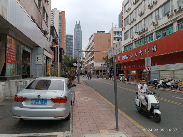 Ein Mann fährt auf einem Scooter eine Straße mit hohen Gebäuden entlang, mit Menschen auf den Gehwegen und Fahrzeugen auf der Straße, unter einem klaren blauen Himmel mit Bäumen im Hintergrund.