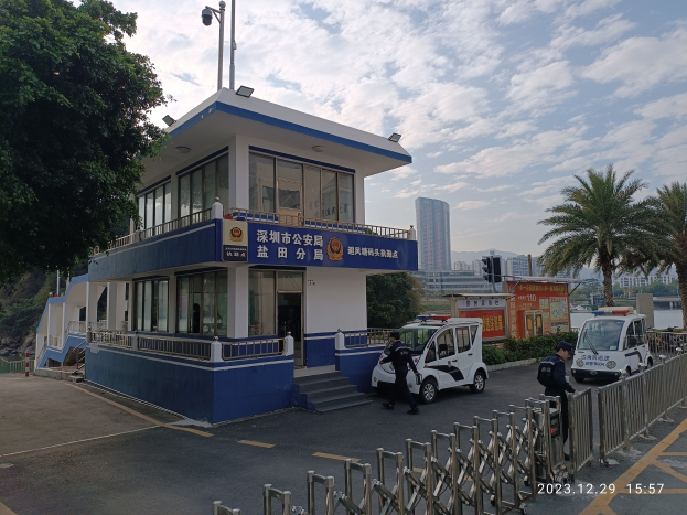 Blau-weißes Gebäude mit der Aufschrift 'Hong Kong Police Station' und einem vor dem Gebäude geparkten Polizeiwagen, Fahrzeuge auf der Straße, Fußgänger, ein Geländer, Bäume, andere Gebäude und eine bewölkte Himmel im Hintergrund.