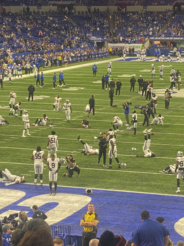 Ein Fußballspiel wird in einem Stadion mit Zuschauern und einigen Sitzenden gespielt, während eine Person eine Kamera hält, was auf eine Live-Übertragung hinweist.