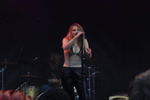 Frau singt in ein Mikrofon auf einer Bühne mit einem Schlagzeuger hinter ihr und einem Publikum vor ihr unter dramatischer Beleuchtung.