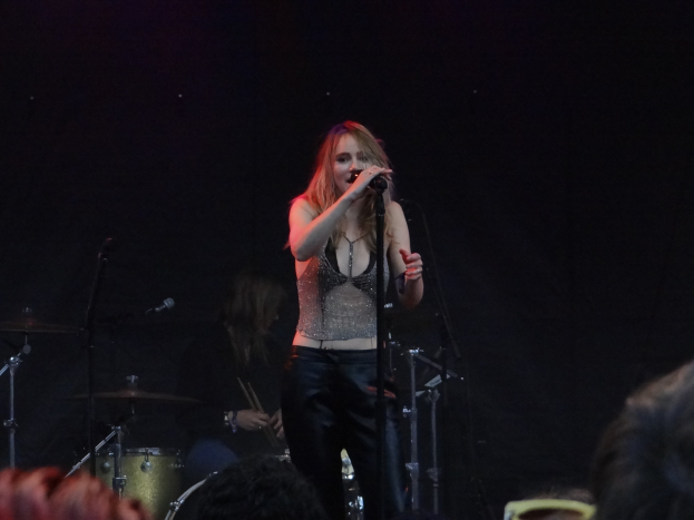 Frau singt in ein Mikrofon auf einer Bühne mit einem Schlagzeuger hinter ihr und einem Publikum vor ihr unter dramatischer Beleuchtung.