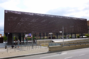 Gro├čes modernes Geb├Ąude mit zahlreichen Fenstern, das als neuer Eingang zur Stadtmitte dient, flankiert von Stra├čeninfrastruktur und Passanten.