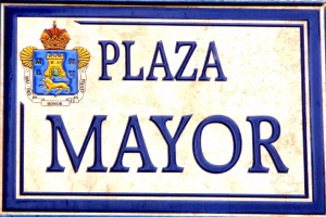Plaque auf einem Gebäude mit der Aufschrift "Plaza Mayor" mit einem Wappen-Logo mit einer Krone, vor einem weißen Hintergrund mit einem blauen Rand und schwarzer fetter Schrift.