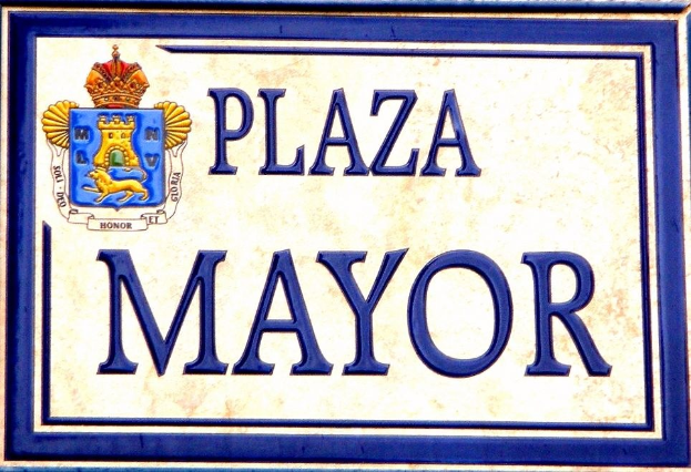 Plaque auf einem Gebäude mit der Aufschrift "Plaza Mayor" mit einem Wappen-Logo mit einer Krone, vor einem weißen Hintergrund mit einem blauen Rand und schwarzer fetter Schrift.