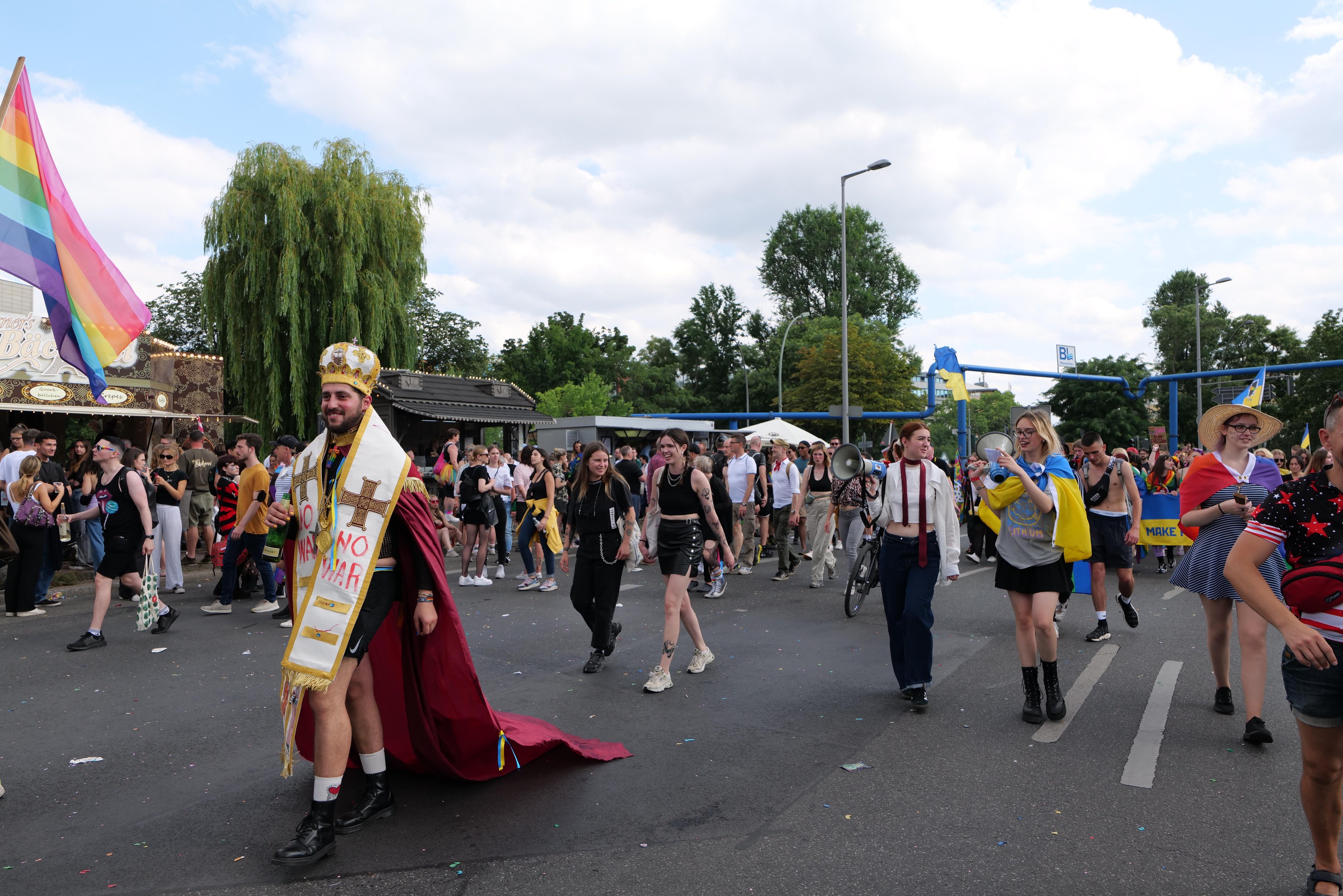 Eine Gruppe von Menschen, die bei der Gay Pride Parade 2018 marschieren und eine Regenbogenflagge sowie Musikinstrumente tragen, während einige eine Mütze tragen und im Hintergrund Laternenpfähle, Bäume, Hütten und ein bewölkter Himmel zu sehen sind.