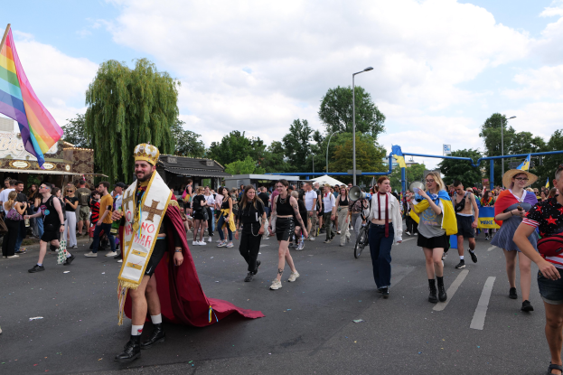 Eine Gruppe von Menschen, die bei der Gay Pride Parade 2018 marschieren und eine Regenbogenflagge sowie Musikinstrumente tragen, während einige eine Mütze tragen und im Hintergrund Laternenpfähle, Bäume, Hütten und ein bewölkter Himmel zu sehen sind.