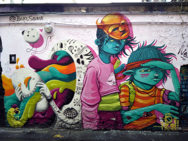 Mural an einer Gebäudeseite mit Graffiti, darunter eine Straße, Strommasten und einen klaren blauen Himmel im Hintergrund.