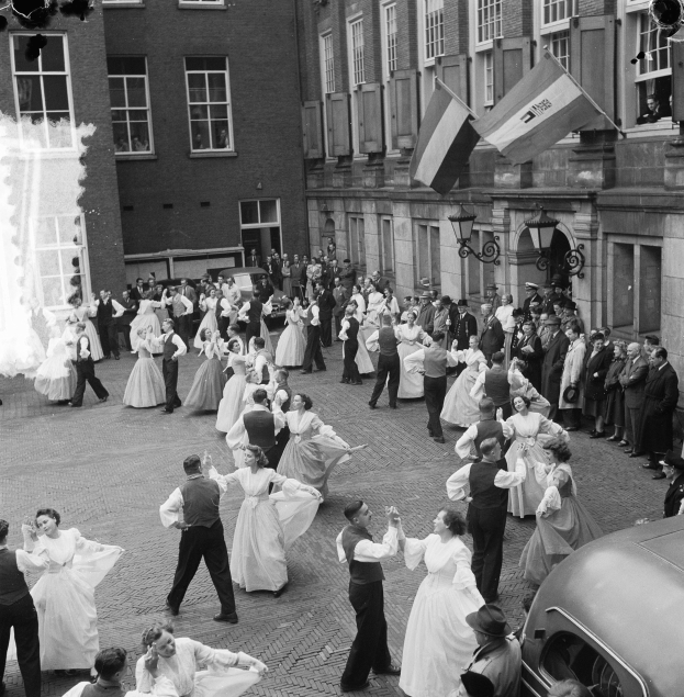 Eine Gruppe von Menschen, die auf der Straße tanzen, vor einem Gebäude mit einem Auto auf der rechten Seite, Flaggen und Lichter an der Wand des Gebäudes im Hintergrund, in Schwarz-Weiß dargestellt.