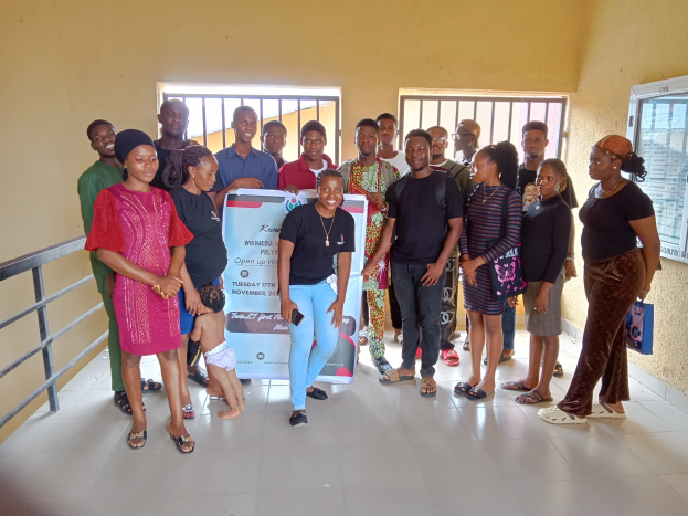 Eine Gruppe von Menschen, die zusammen in einem Raum stehen und eine Tafel in der Mitte halten, mit einem Geländer auf der linken Seite, Fenstern und einer Wand im Hintergrund.