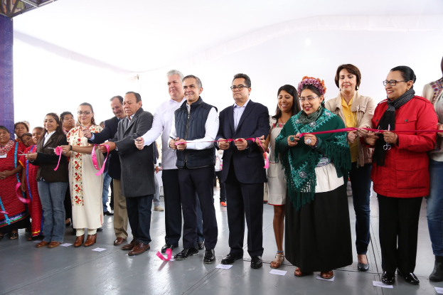 Eine Gruppe von Menschen bei einer Eröffnungszeremonie mit einer Schere, die vor einer Menge lächeln, mit einer Säule links, verstreuten Papieren auf dem Boden und einem weißen Tuch im Hintergrund.