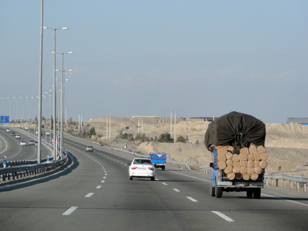 Lastwagen mit einer großen Ladung Holz auf einer Autobahn, umgeben von Geländern, Laternen, Schildern, Bäumen und Sand, mit Hügeln und einem klaren blauen Himmel im Hintergrund.