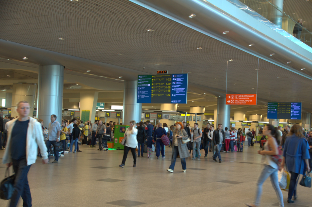 Menschen gehen durch einen Flughafen mit Überhangschildern, Säulen, Deckenlampen und einer Glasbrüstung auf der rechten Seite.