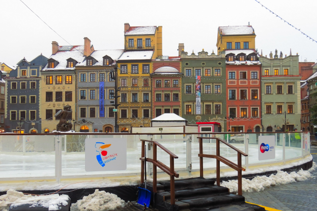 Eislaufbahn in einem Stadtplatz umgeben von Gebäuden, Treppen, Geländern, Informationsschildern, schneebedeckter Fläche, Laternen, Bäumen und Himmel.