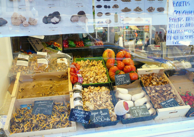 Marktstand mit Auslagevitrine voller verschiedener Lebensmittel wie Gemüse in Schalen, Flaschen und andere Gegenstände, mit textbedruckten Papieren auf dem Glas und ein paar Menschen im Hintergrund.