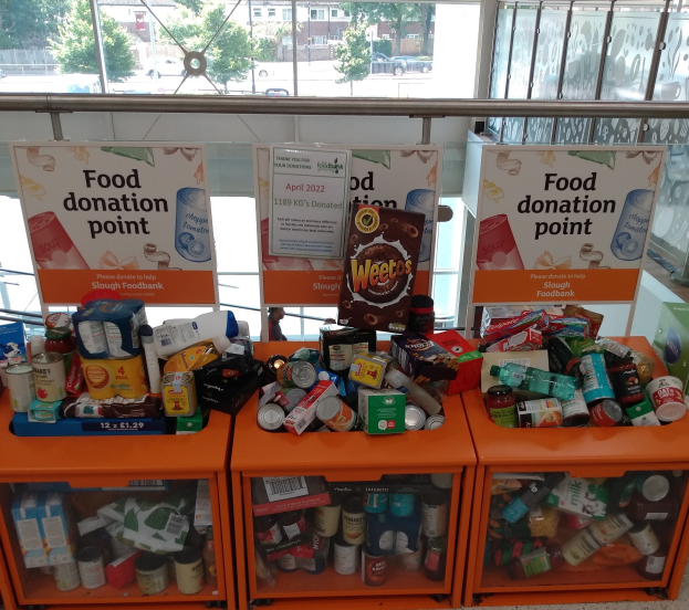 Orange Schübe mit Lebensmittelspenden, darunter Flaschen, Dosen und Kartons, mit informativen Schildern; vor dem Glasfenster sind Fahrzeuge, Bäume, Gebäude und Himmel zu sehen.