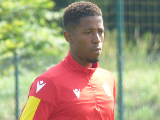 Ein junger Mann in einem roten und gelben T-Shirt steht auf einem Fußballfeld, trainiert mit entschlossener Miene, umgeben von einem Zaun und Bäumen.