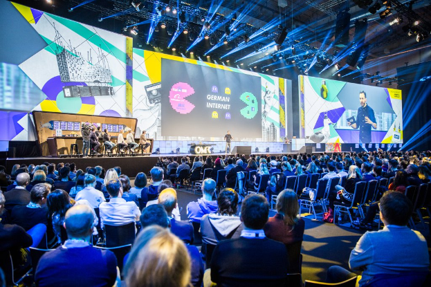 Eine große Menschenmenge, die auf Stühlen vor einer Bühne sitzt, mit einigen Personen, die auf dem Podium stehen, einem Tisch mit verschiedenen Gegenständen links und Bildschirmen und Lichtern im Hintergrund auf der Deutschen Internet-Konferenz 2019.