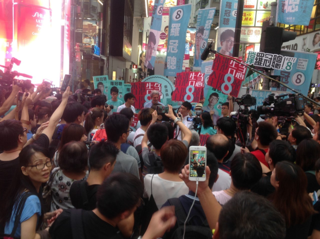 Eine Menschenmenge mit Schildern, Kameras und Mobiltelefonen auf einer belebten Straße mit Gebäuden, Schildern, einer Straßenlaterne und einer Ampel im Hintergrund unter einem bewölkten Himmel.