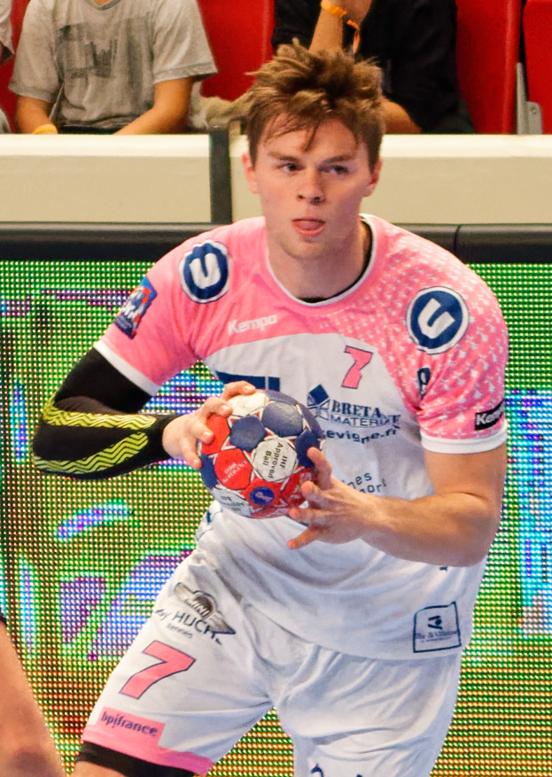Ein Mann in einem rosa Hemd, der einen Füßball hält, mit einer anderen Person Hand auf der linken Seite und ein paar Menschen im Hintergrund auf Stühlen sitzend.