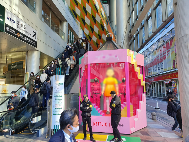 Eine Gruppe von Menschen steht vor einem gläsernen Gebäude mit Säulen und Texttafeln, einige tragen Masken, während eine Rolltreppe mit Menschen zu sehen ist. In der Mitte steht ein pinker Kasten mit Schaufensterpuppen und der Aufschrift "Netflix' neue Netflix-Bühne in Tokio."