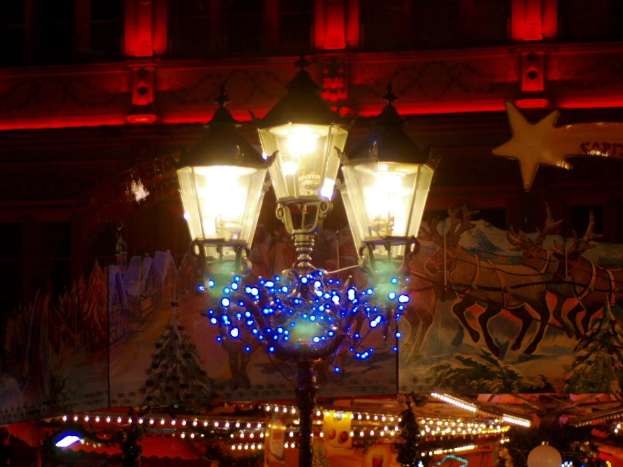 Eine mit Lichtern und Festdekoration geschmückte Straßenlaterne steht nachts in einer Stadt, im Hintergrund ist ein Gebäude zu sehen.