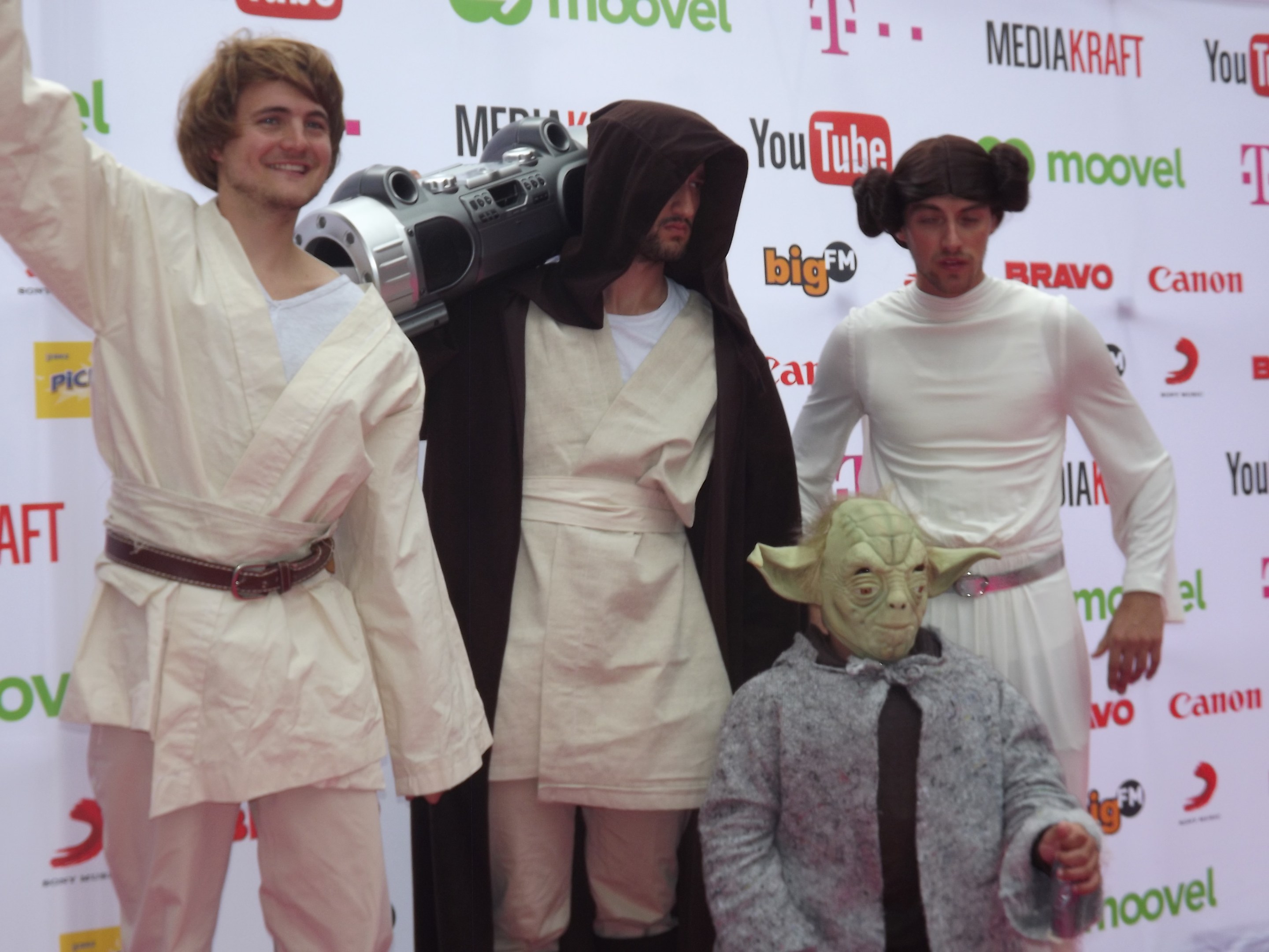 Gruppe von Menschen in Star-Wars-Kostümen auf einem roten Teppich posend, wobei einer ein Objekt in der Mitte hält und ein Banner mit Text und Logos im Hintergrund zu sehen ist.