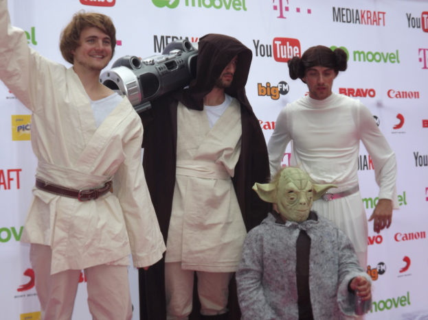 Gruppe von Menschen in Star-Wars-Kostümen auf einem roten Teppich posend, wobei einer ein Objekt in der Mitte hält und ein Banner mit Text und Logos im Hintergrund zu sehen ist.