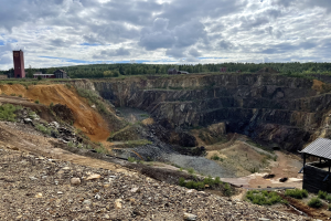 Großer Tagebau auf einem Schmutzfeld umgeben von Bäumen, Gebäuden und einem Schuppen, mit bewölktem Himmel und Boden bedeckt mit Steinen und Pflanzen.