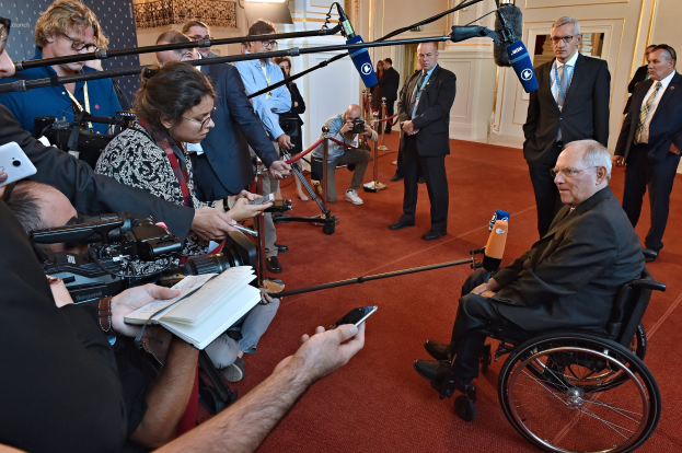 Ein Mann im Rollstuhl ist von Reportern umgeben, die Kameras, Handys und Mikrofone mit Ständern halten, mit einem Banner, Scheinwerfern und einer Wand im Hintergrund.