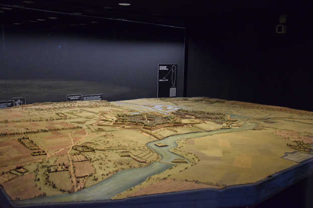 Ein Stadtmodell, das in einem Museum ausgestellt ist, mit einem Tisch im Vordergrund und einer Texttafel im Hintergrund, unter einer Decke mit warmem Licht.