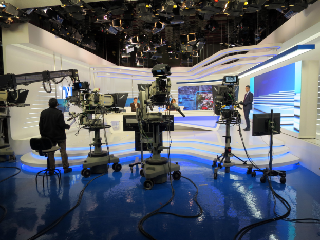 Ein Fernsehstudio mit mehreren Kameras auf dem Boden, einer sitzenden Person im Vordergrund und einigen stehenden Personen im Hintergrund, mit Deckenmontagelampen für eine Livesendung.