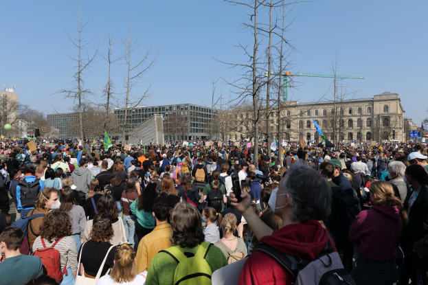 Eine große Menge von Menschen, die an einer Klimawandel-Demonstration vor einem Gebäude mit Fenstern, Bäumen und einem klaren Himmel teilnehmen.