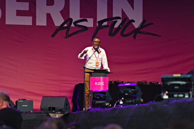 Ein Mann in einem weißen Hemd und braunen Hosen steht auf einer Bühne und hält ein Mikrofon in der Hand, mit einem pinken Banner mit der Aufschrift "Berlin als Fuck" im Hintergrund und einer Menge davor.