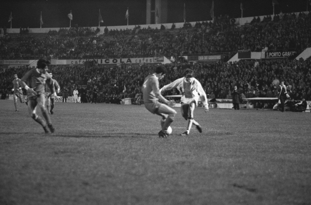 Schwarzes und weißes Bild von Männern, die Fußball in einem Stadion spielen, mit Zuschauern auf den Tribünen und Fahnen im Hintergrund.