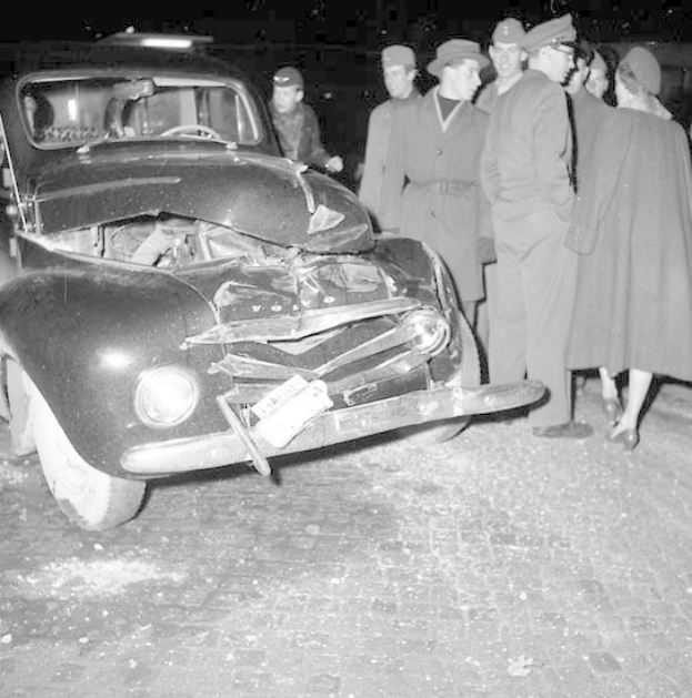 Schwarz-weißes Foto eines stark beschädigten Autos am Straßenrand mit eingedellter Motorhaube und zersplitterten Fenstern, umgeben von einer Gruppe von Menschen, die es inspizieren.