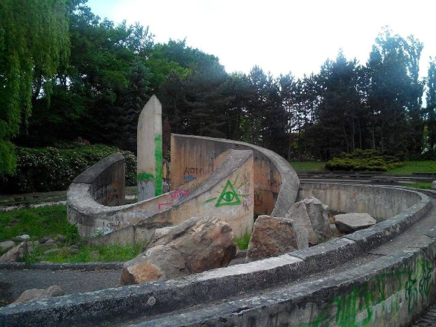 Verlassener Skatepark mit Graffiti an seiner Seite, umgeben von Felsen, Gras, Pflanzen, Bäumen und einem klaren blauen Himmel.