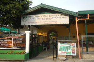 Der Eingang der San Lorenzo Ruiz Pfarrschule zeigt eine Tafel mit Text, einen Metallzaun, Menschen, die unter einem Dach sitzen, Pfosten, ein Schild, Pflanzen, einen Baum und einen bewölkten Himmel.