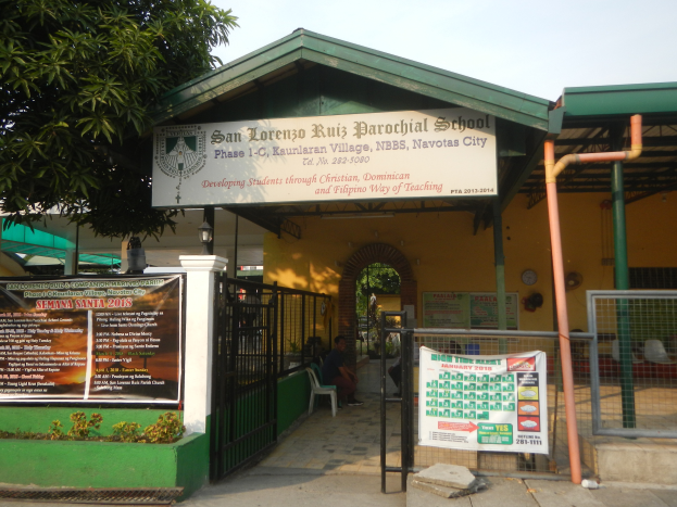 Der Eingang der San Lorenzo Ruiz Pfarrschule zeigt eine Tafel mit Text, einen Metallzaun, Menschen, die unter einem Dach sitzen, Pfosten, ein Schild, Pflanzen, einen Baum und einen bewölkten Himmel.