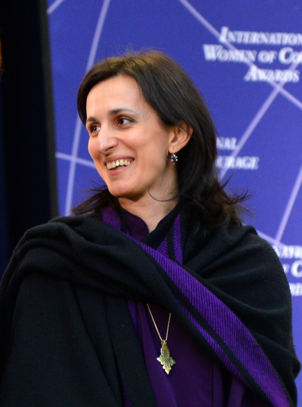 Eine Frau in einem lila Kleid und schwarzer Stola steht neben einer anderen Frau, beide lächeln, mit einem Banner im Hintergrund, auf dem "Internationale Frauen der Farbe" steht.