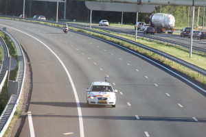 Ein Auto fährt auf einer Autobahn mit einem Polizeiwagen am Straßenrand, anderen Fahrzeugen, Geländern, Pfählen, Planken und Bäumen im Hintergrund.