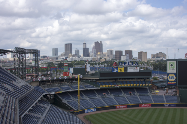 Ein Baseballstadion mit leeren Sitzplätzen, Pfosten und Beschilderung, vor einem bewölkten Himmel und einer fernen Stadtkulisse mit Bäumen und Gebäuden.