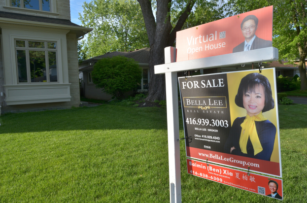 Ein Immobilien-Verkaufsschild steht vor einem Haus mit Fenstern, umgeben von grünem Gras, Bäumen und Pflanzen, unter einem sichtbaren Himmel.