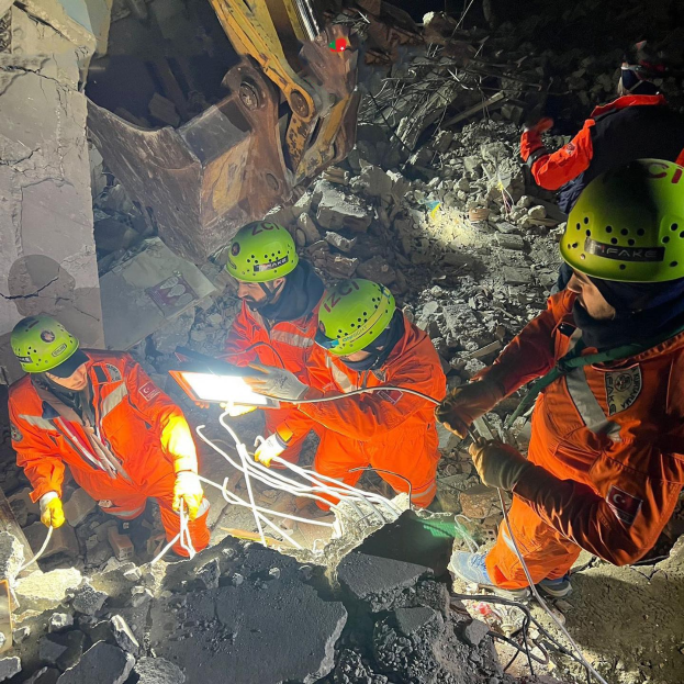 Eine Gruppe von Menschen in orangefarbenen Uniformen und grünen Helmen bei der Arbeit auf Schutt mit einem Kran im Hintergrund bei einer Rettungsaktion.