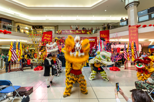 Ein lebendiges chinesisches Neujahrsfest in einem Einkaufszentrum mit Menschen in Kostümen, die tanzen, anderen, die auf Stühlen sitzen und Handys halten, mit Flaggen, Bannern, Dekorationen, Geländern, Säulen und Deckenlampen im Hintergrund.