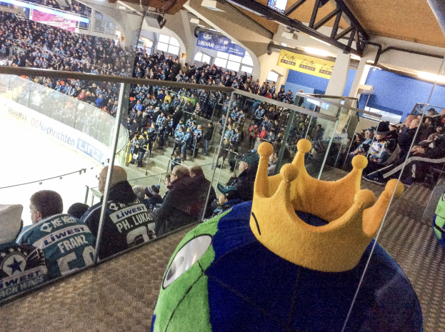 Menge Menschen in einer Arena bei einem Eishockeyspiel mit einer Person mit Krone im Vordergrund, eine Glaswand dahinter, Banner, Säulen und Deckenlampen.