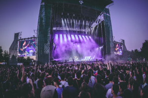 Eine große Menschenmenge auf einem Musikfestival mit einer Bühne, auf der Musiker spielen, beleuchtet von Lichtern und umgeben von Bildschirmen, vor dem Hintergrund eines Gebäudes, Bäume und eines klaren blauen Himmels.