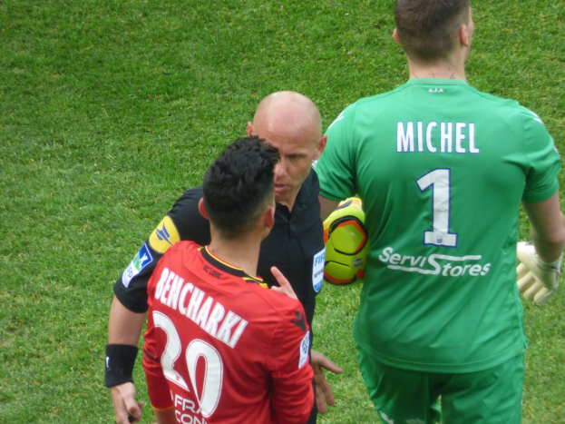 Schiedsrichter spricht mit zwei Füßballspielern auf einem grünen Rasen, ein Spieler hält einen Ball.