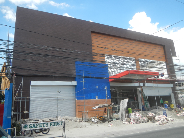 Baustelle mit einem "Sicherheit geht vor"-Schild am Gebäude, Fahrzeuge auf der Straße, eine Texttafel, Menschen in der Nähe, Bäume, Versorgungsmasten, Drähte und Himmel im Hintergrund.