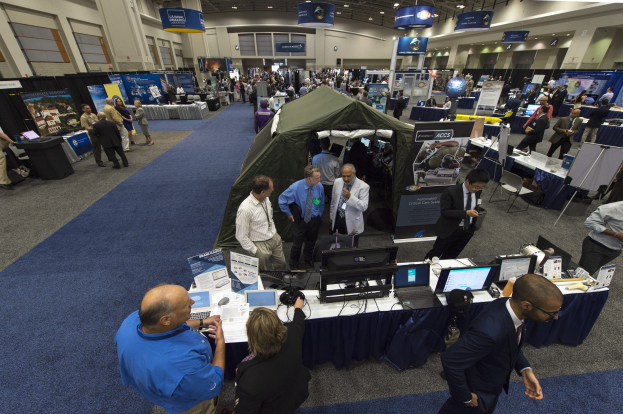 Eine große Gruppe von Menschen auf einer Karriere-Messe um einen Tisch mit Laptops, Monitoren und Papieren versammelt, mit einem Zelt, Informations-Boards, Deckenleuchten und Fenstern im Hintergrund.
