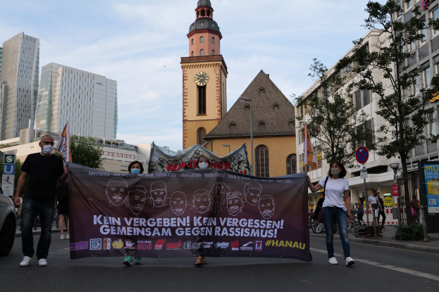 Eine Gruppe von Menschen in Masken, die eine Straße entlanggehen und ein Banner mit Text und Bildern halten, mit einem geparkten Auto auf der linken Seite, Gebäuden und Bäumen im Hintergrund und einem Kirchturm unter einem klaren blauen Himmel.